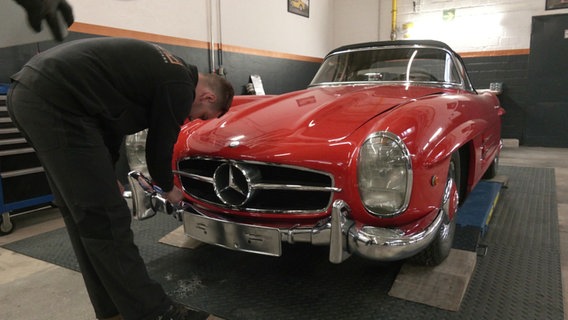 Ein Mann in schwarzer Kleidung steht in einer Werkstatt. Mit einem Werkzeug geht er an einen roten Mercedes 300 SL Roadster aus den 1950er Jahren ran. Die Stoßstangen und der Grill glänzen in Chromfarben. © Screenshot 