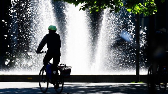 Ein Fahrradfahrer radelt bei Sonnenschein vor einem Brunnen auf dem Schlossplatz. © dpa Foto: Julian Stratenschulte